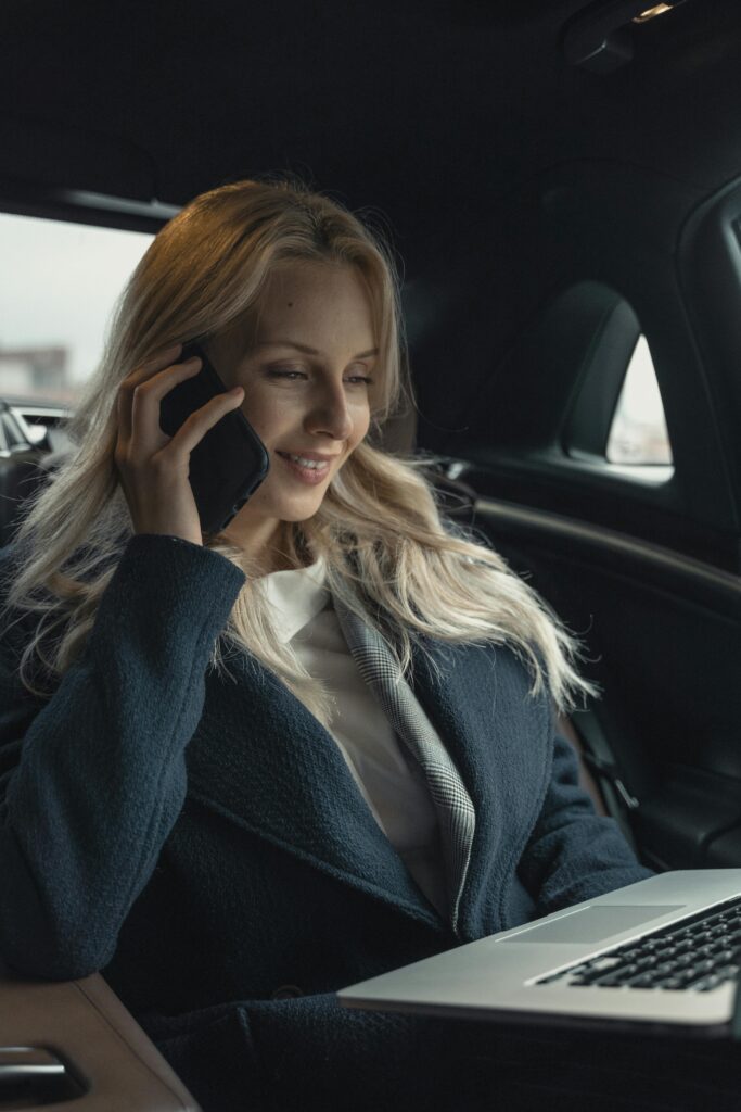 Professional woman in a car, talking on mobile phone, with laptop open, working remotely.